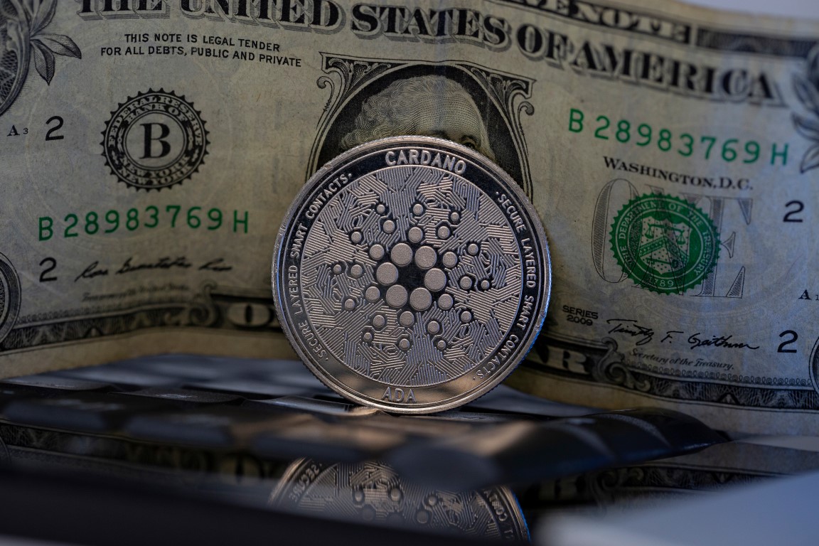 Picture of a Cardano coin standing in front of a U.S. dollar bill