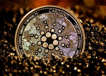Picture of a gold and black Cardano coin half-sunk into the sand