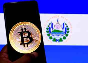 Picture of a Bitcoin logo on a smartphone in front of a flag of El Salvador