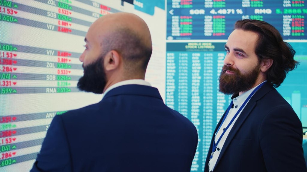 رجلان ببدلات رسمية ينظران إلى شاشة تعرض بيانات السوق المالي، بما في ذلك أسعار الأسهم أو العملات الرقمية